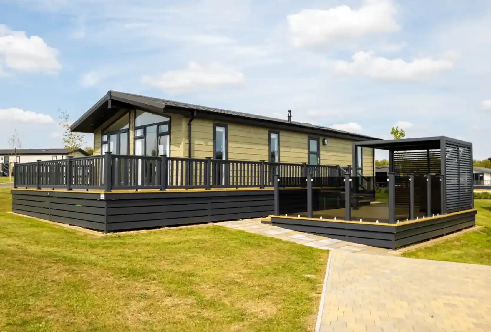 Modern light-green mobile home with a large wooden deck and black railings, adjacent to a covered outdoor seating area.