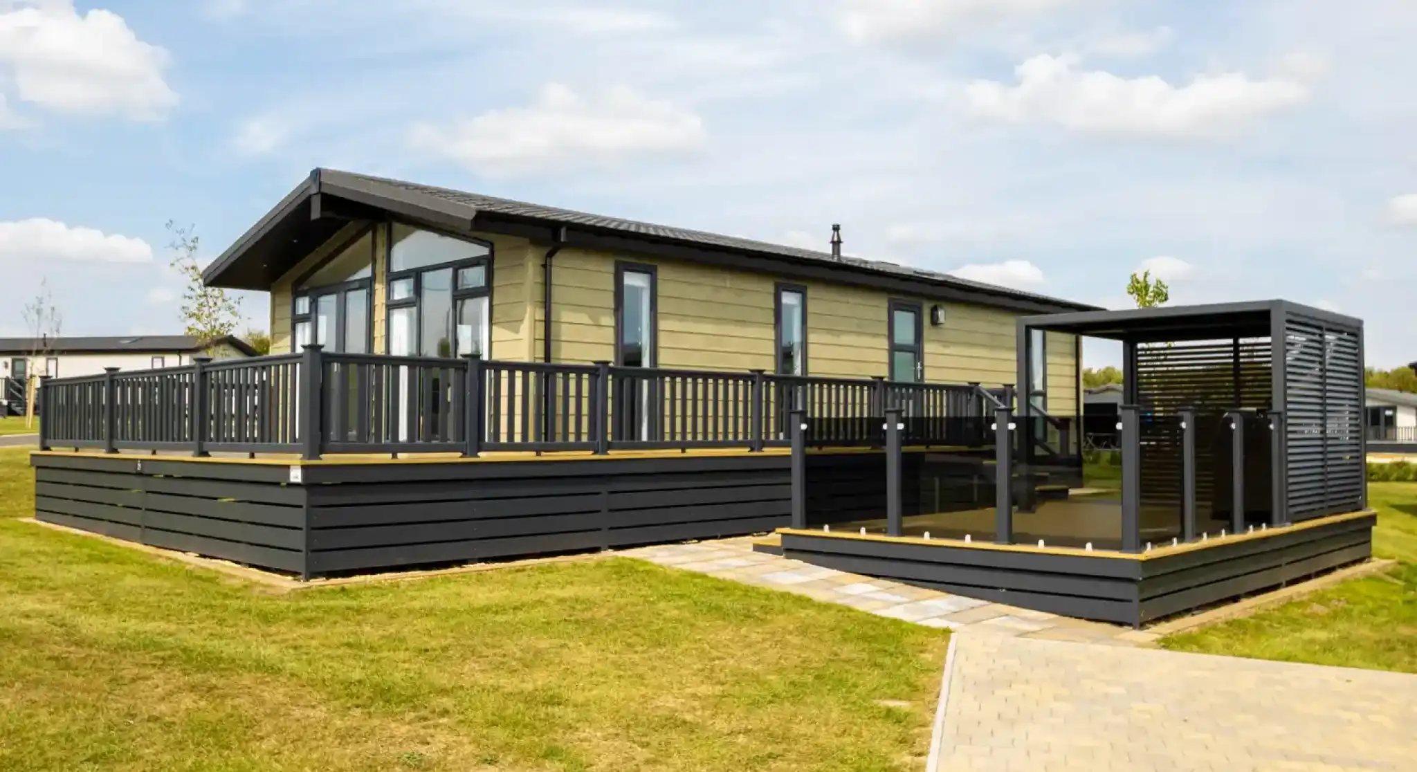Modern light-green mobile home with a large wooden deck and black railings, adjacent to a covered outdoor seating area.