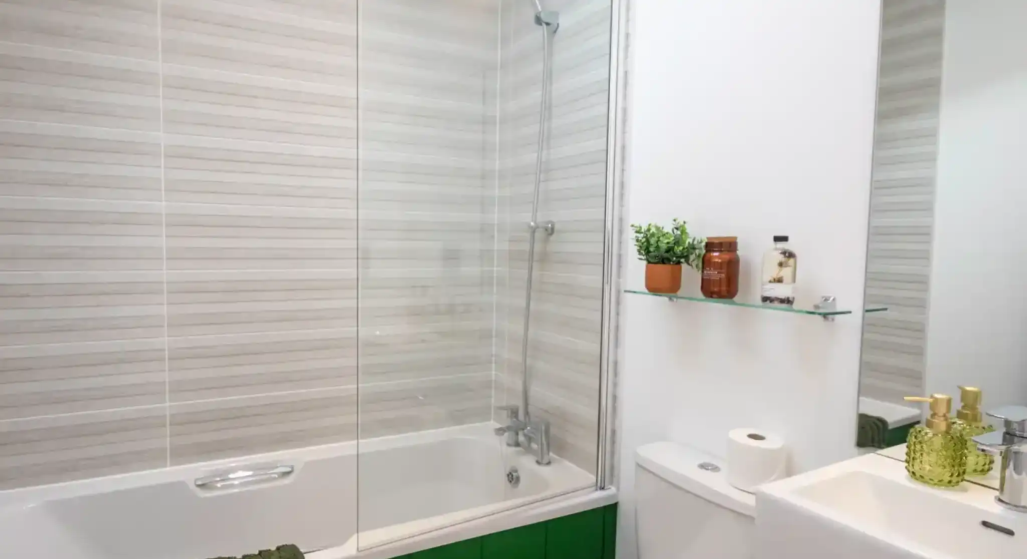 Modern bathroom with a bathtub and glass screen, toilet, sink, green panelling, striped wall tiles, and toiletries on a glass shelf.