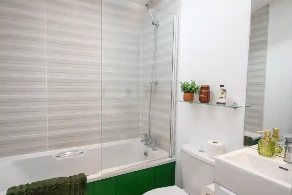 Modern bathroom with a bathtub and glass screen, toilet, sink, green panelling, striped wall tiles, and toiletries on a glass shelf.