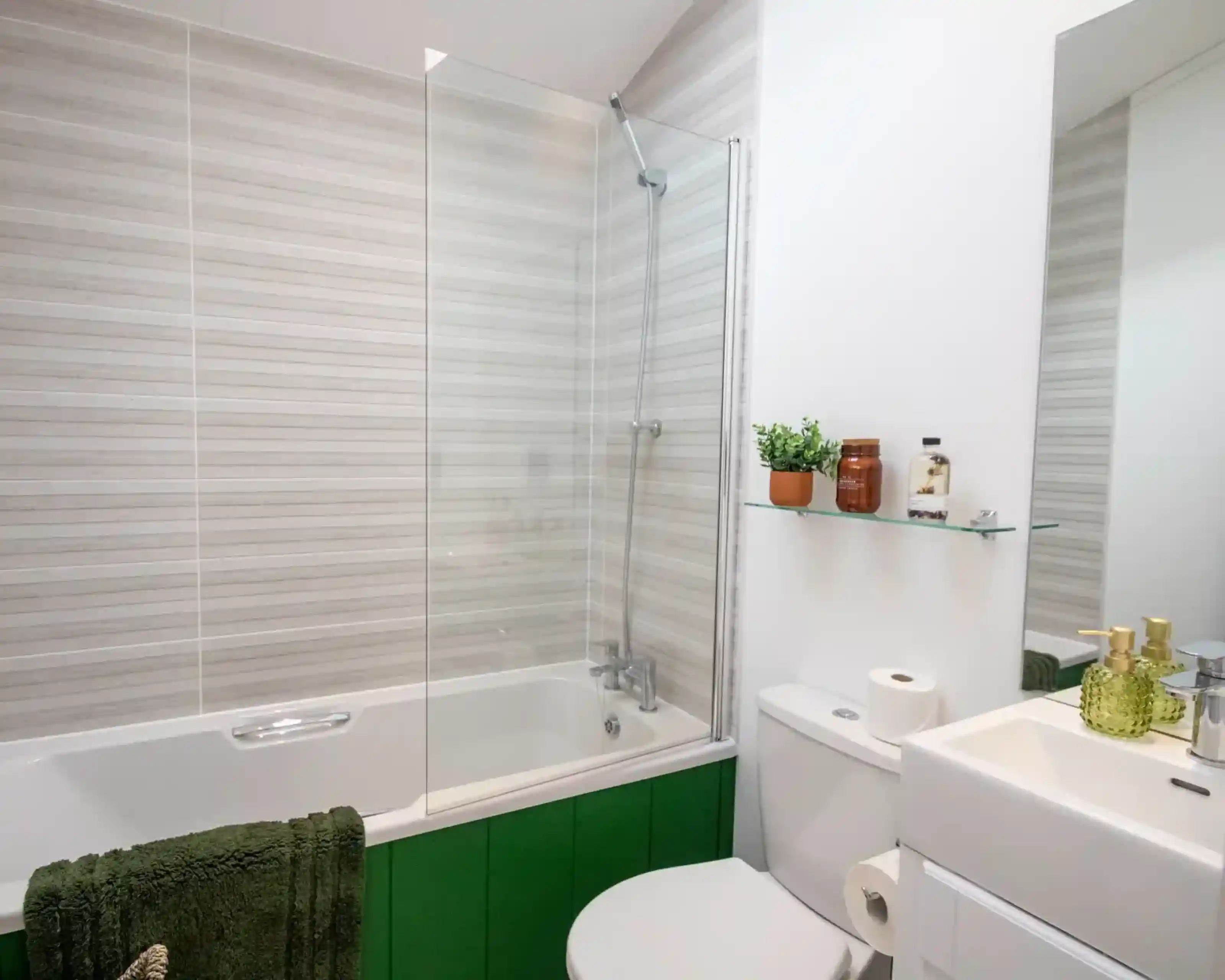 Modern bathroom with a bathtub and glass screen, toilet, sink, green panelling, striped wall tiles, and toiletries on a glass shelf.