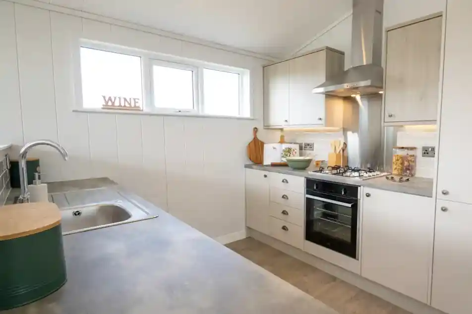 A modern kitchen featuring a stainless steel sink, oven, and range hood with white panelled walls and light wood cabinets.
