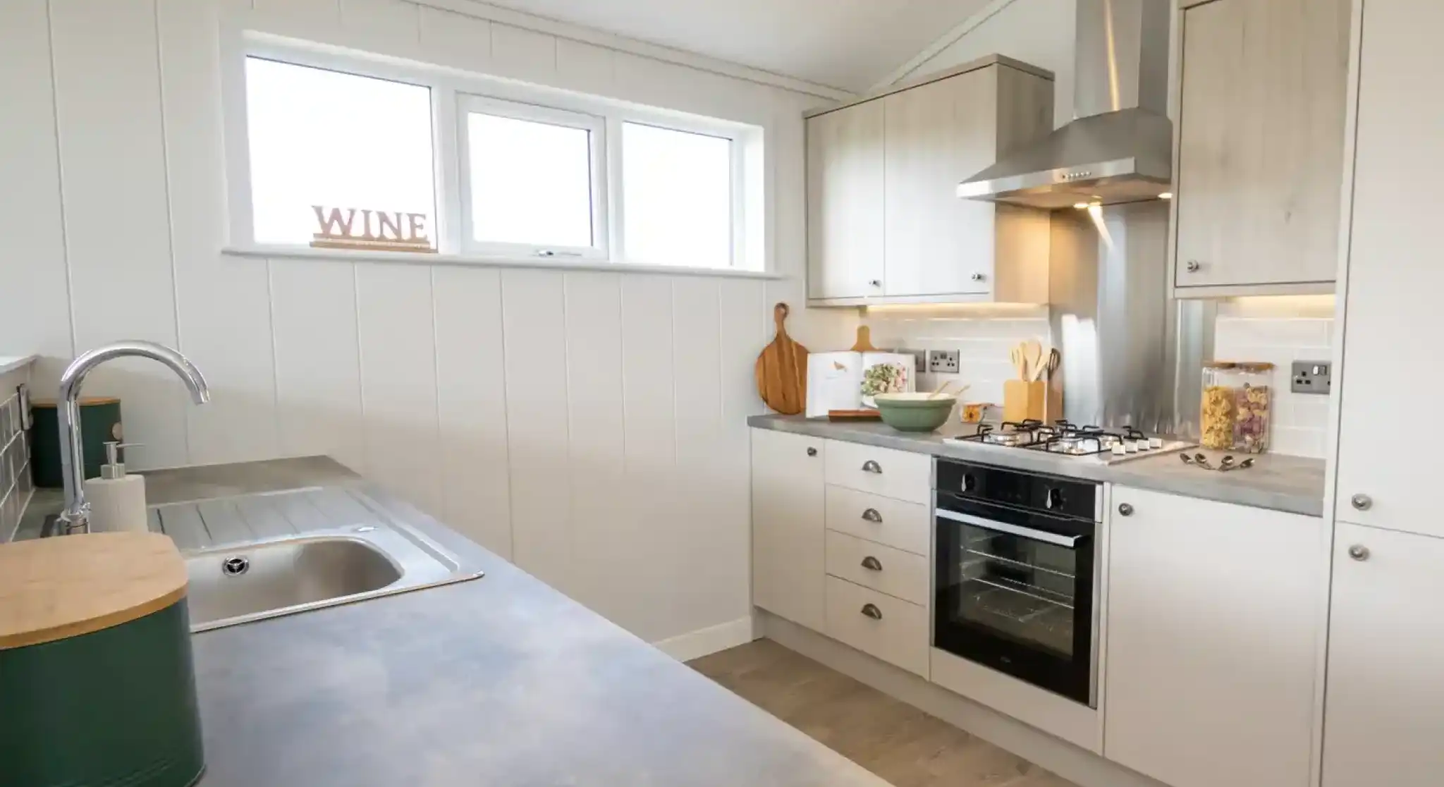 A modern kitchen featuring a stainless steel sink, oven, and range hood with white panelled walls and light wood cabinets.