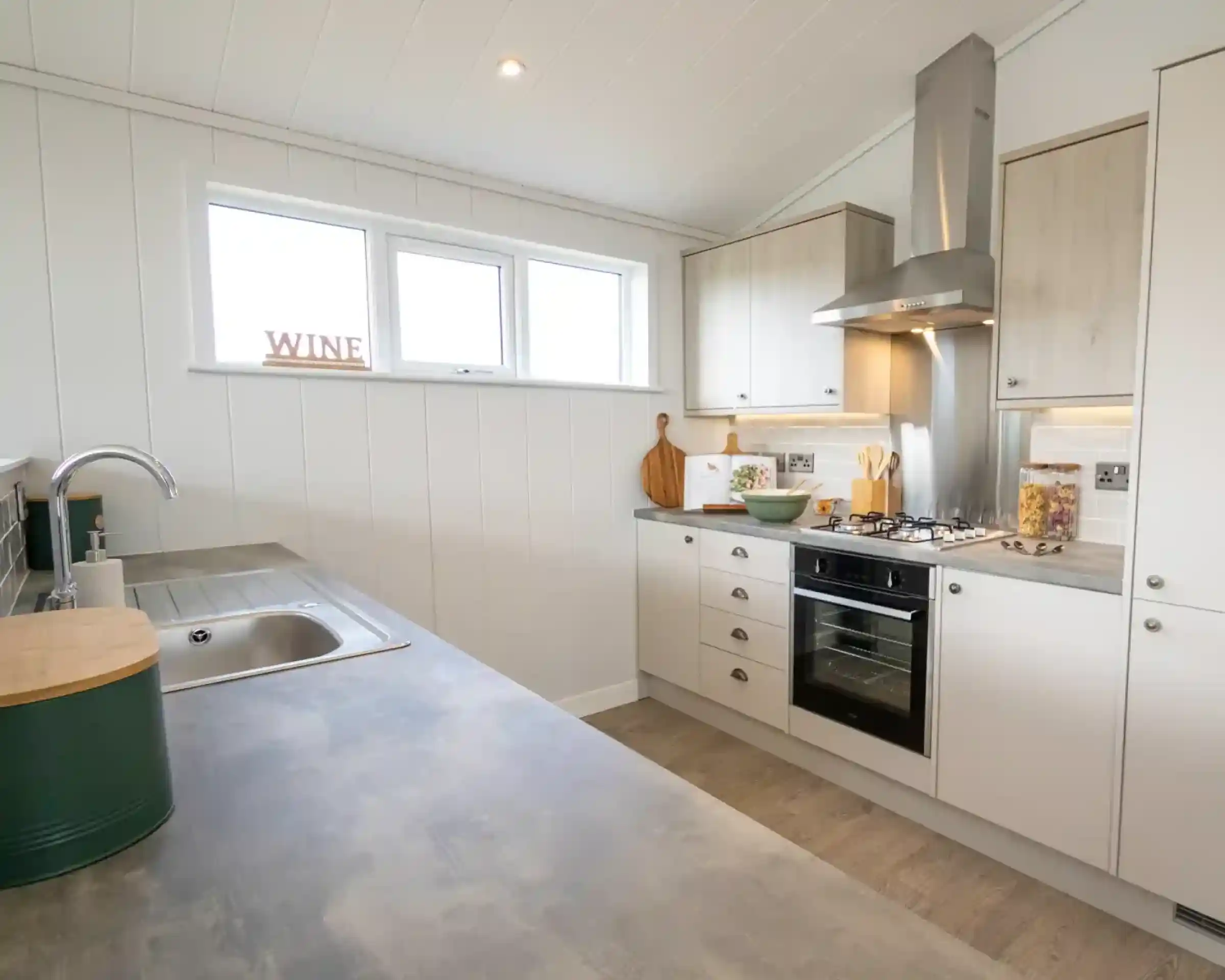 A modern kitchen featuring a stainless steel sink, oven, and range hood with white panelled walls and light wood cabinets.