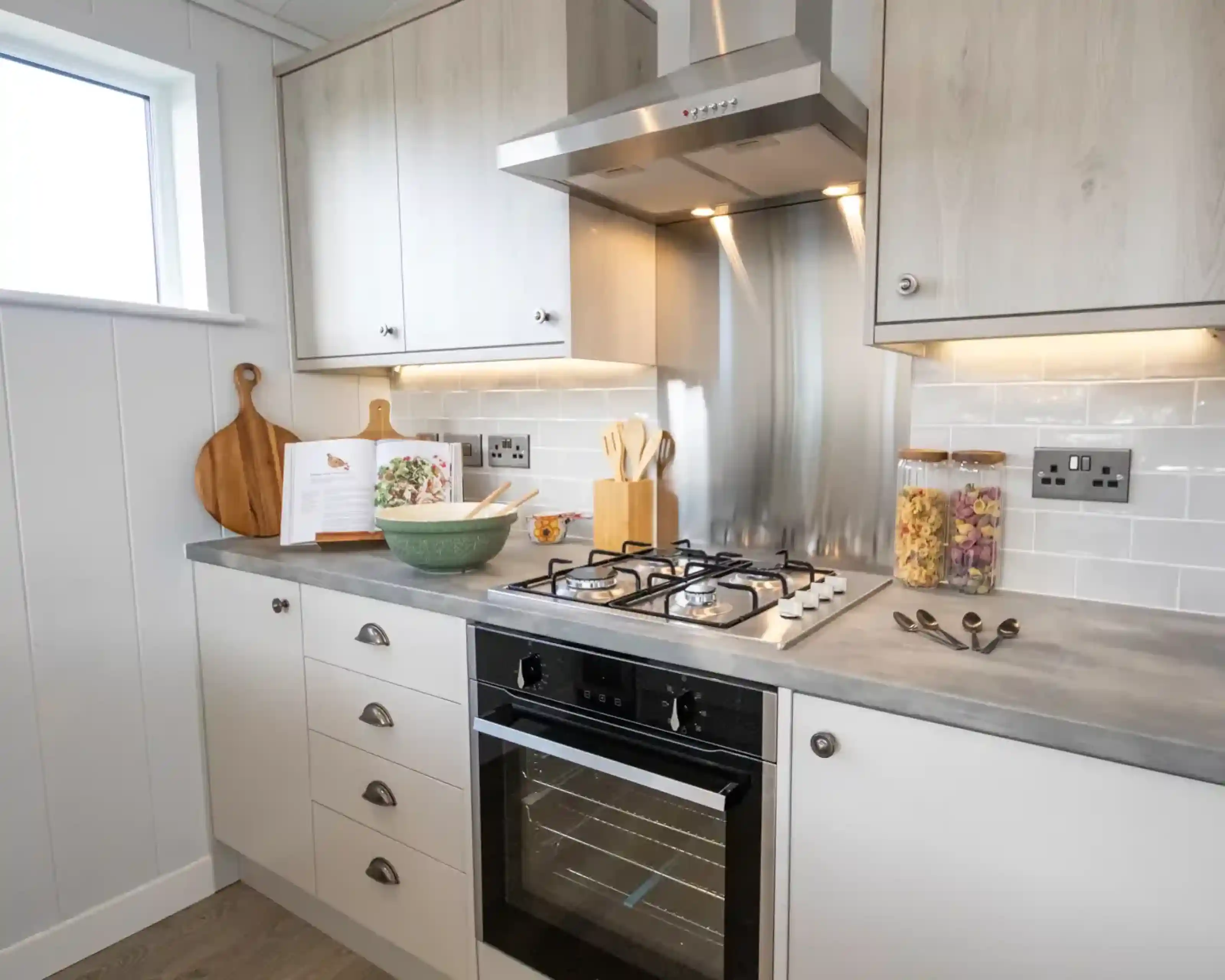 A modern kitchen counter features a gas stove, oven, cookbook, mixing bowl, and decorative jars of pasta.
