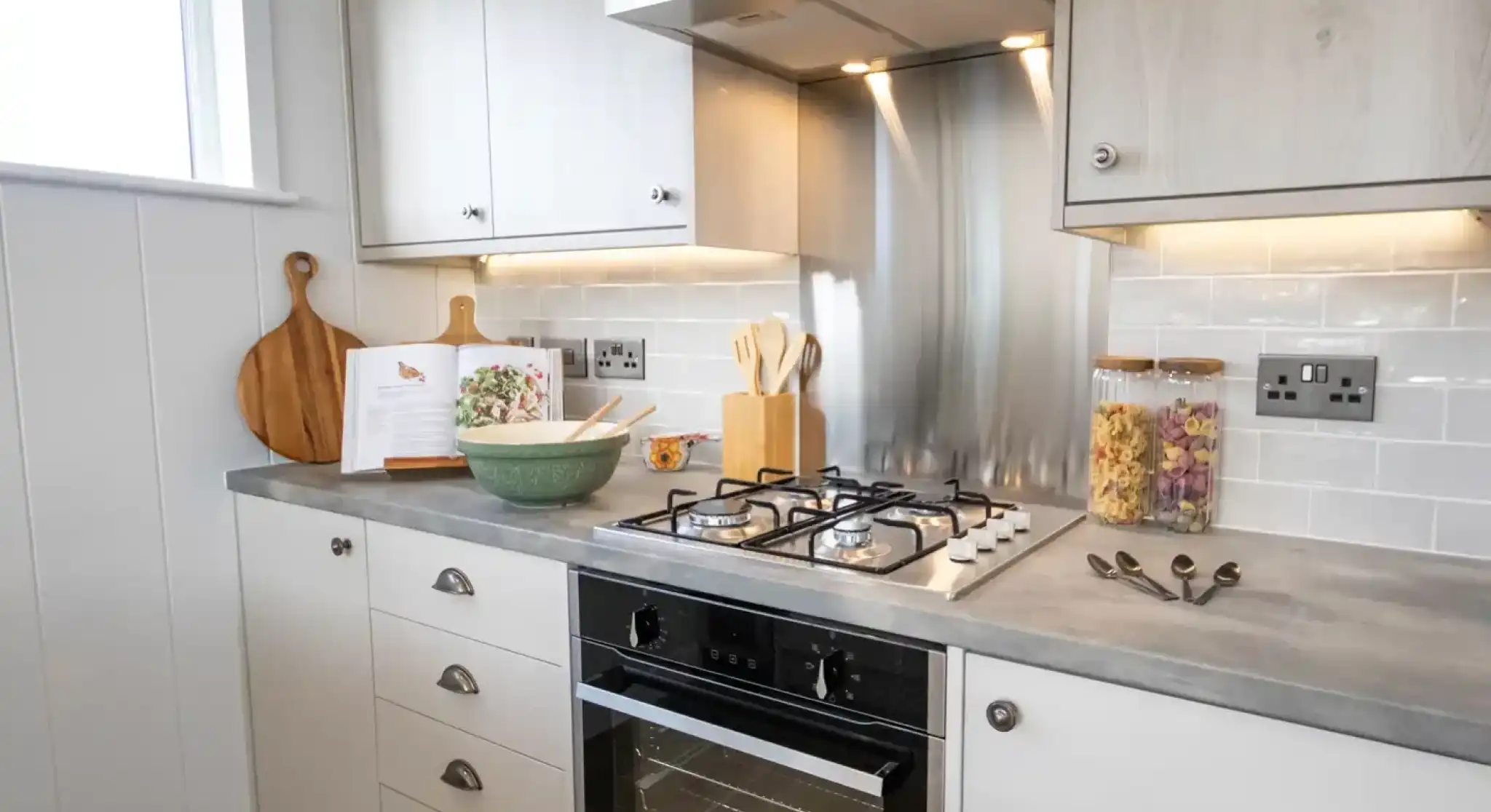 A modern kitchen counter features a gas stove, oven, cookbook, mixing bowl, and decorative jars of pasta.