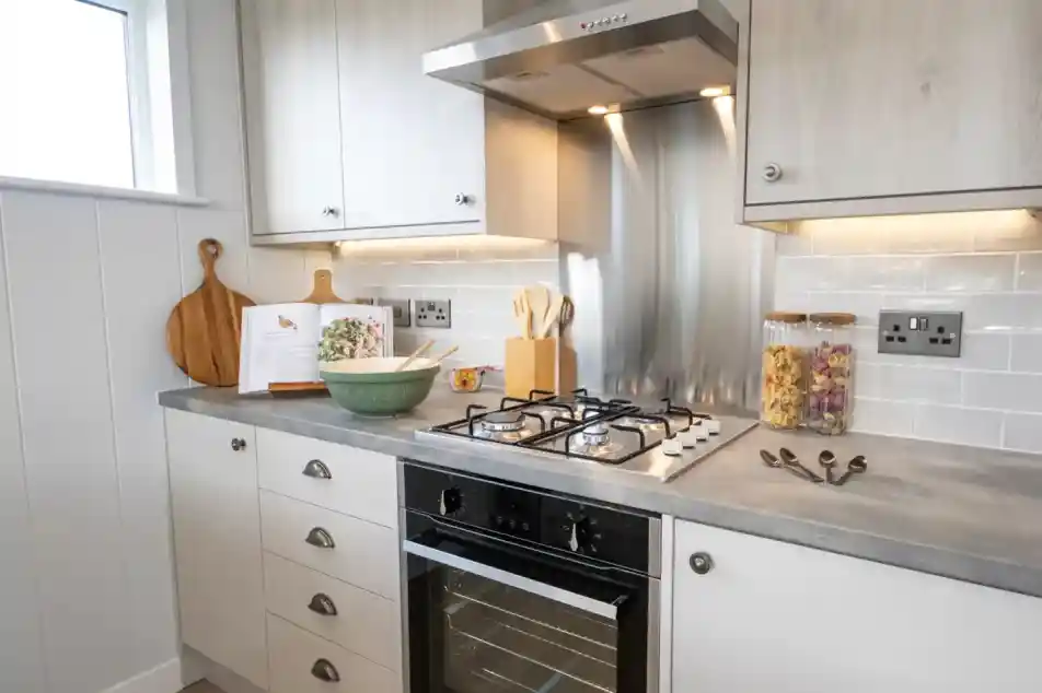 A modern kitchen counter features a gas stove, oven, cookbook, mixing bowl, and decorative jars of pasta.