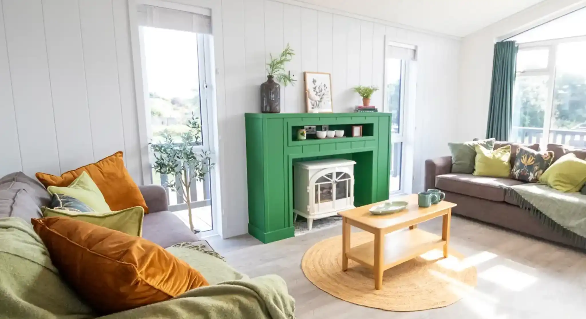 A cozy living room with a green fireplace as the centerpiece. Gray sofas adorned with colorful cushions are arranged around a wooden coffee table. Natural light streams in through large windows, illuminating the space. A round jute rug adds warmth to the floor, and potted plants are placed decoratively on the mantel.