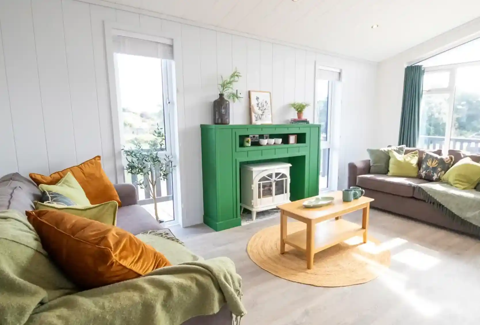 A cozy living room with a green fireplace as the centerpiece. Gray sofas adorned with colorful cushions are arranged around a wooden coffee table. Natural light streams in through large windows, illuminating the space. A round jute rug adds warmth to the floor, and potted plants are placed decoratively on the mantel.