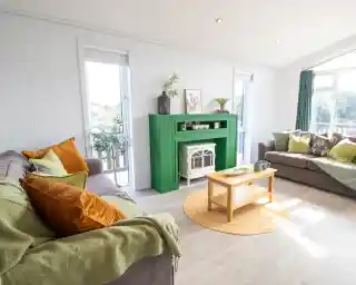 A cozy living room with a green fireplace as the centerpiece. Gray sofas adorned with colorful cushions are arranged around a wooden coffee table. Natural light streams in through large windows, illuminating the space. A round jute rug adds warmth to the floor, and potted plants are placed decoratively on the mantel.