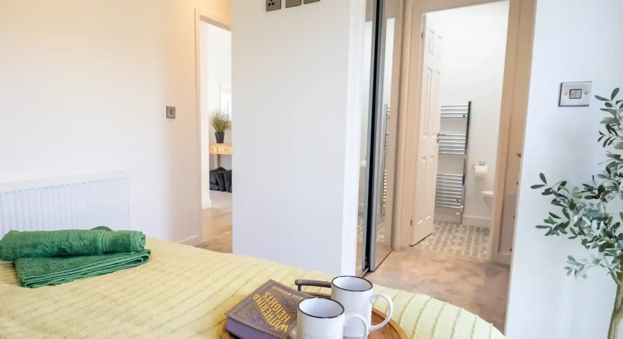 A bed with a yellow textured blanket and green towels features a wooden tray with a book and two mugs, with an open doorway to a bathroom in the background.