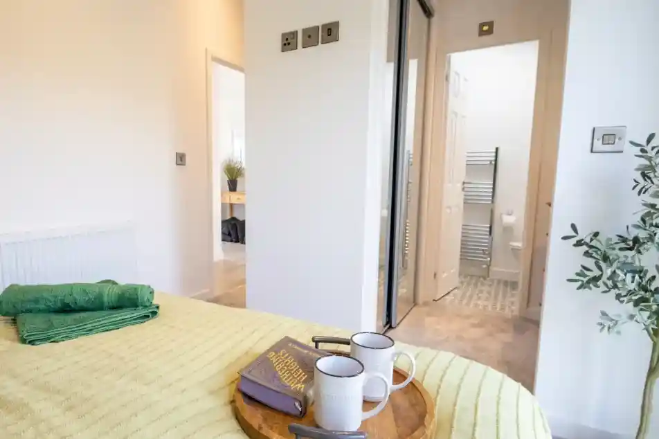 A bed with a yellow textured blanket and green towels features a wooden tray with a book and two mugs, with an open doorway to a bathroom in the background.
