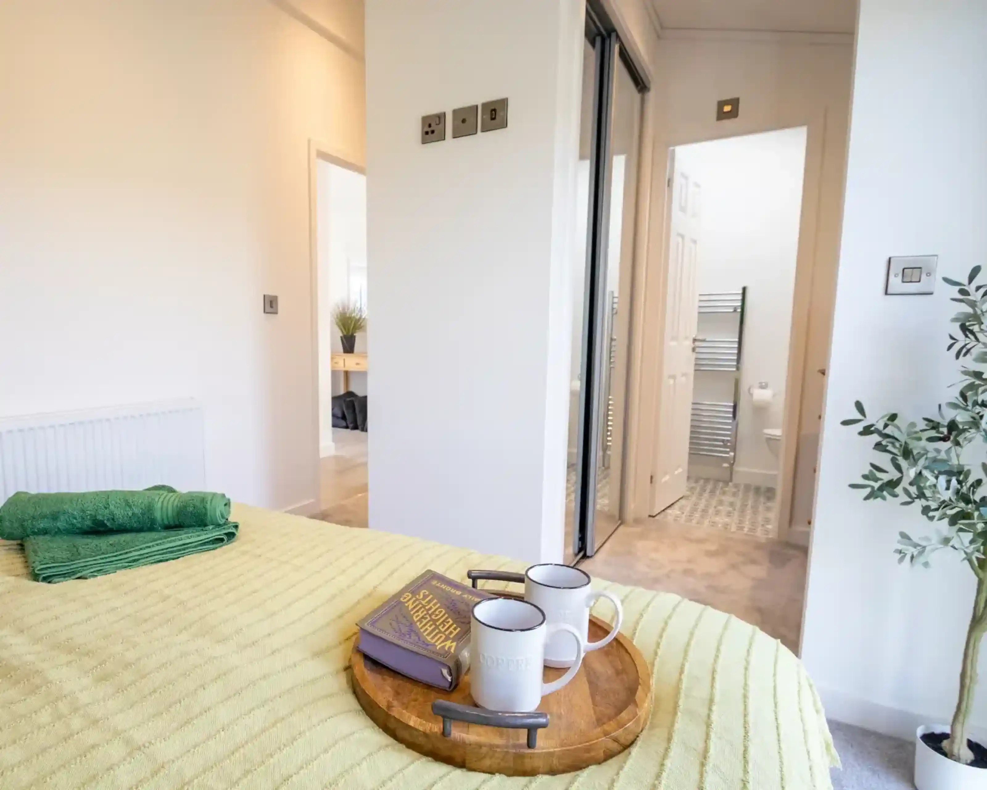 A bed with a yellow textured blanket and green towels features a wooden tray with a book and two mugs, with an open doorway to a bathroom in the background.