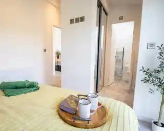 A bed with a yellow textured blanket and green towels features a wooden tray with a book and two mugs, with an open doorway to a bathroom in the background.