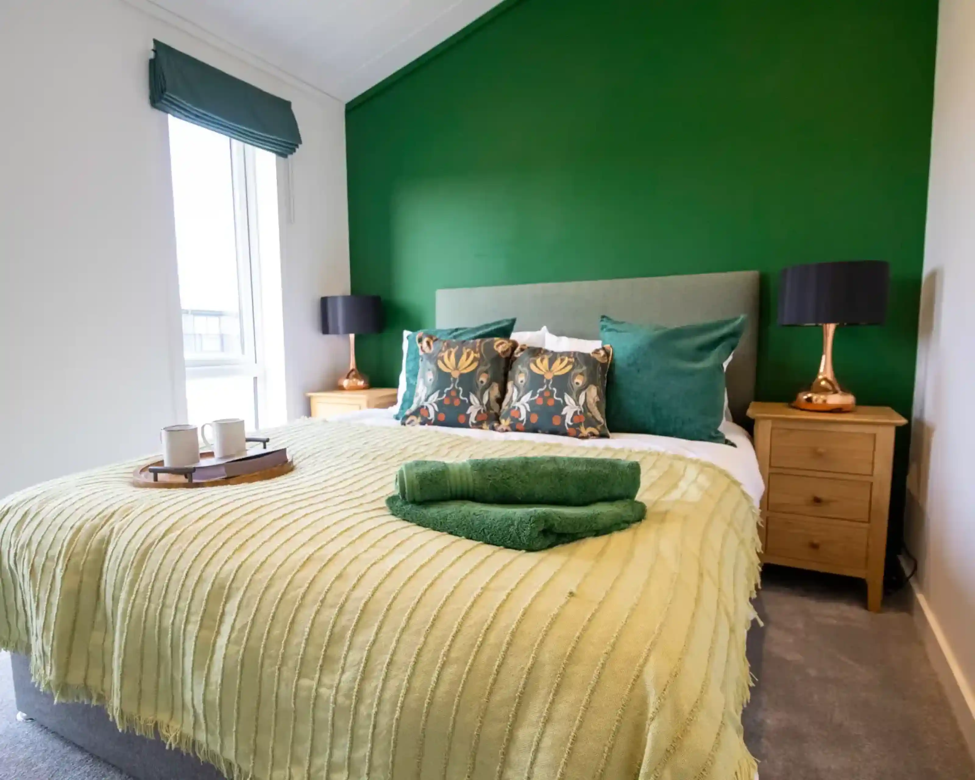 A brightly decorated bedroom with a large green accent wall behind a bed with green and patterned pillows, a light green blanket, and a tray with two mugs.