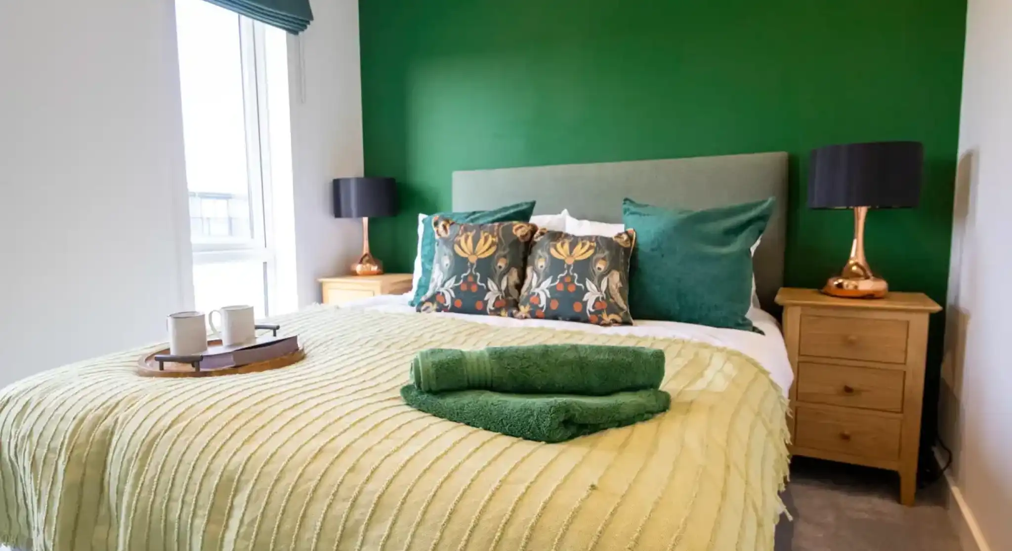 A brightly decorated bedroom with a large green accent wall behind a bed with green and patterned pillows, a light green blanket, and a tray with two mugs.