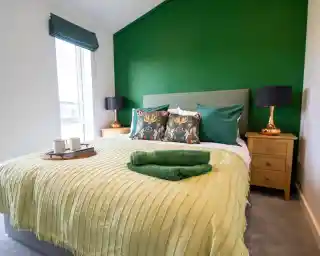 A brightly decorated bedroom with a large green accent wall behind a bed with green and patterned pillows, a light green blanket, and a tray with two mugs.