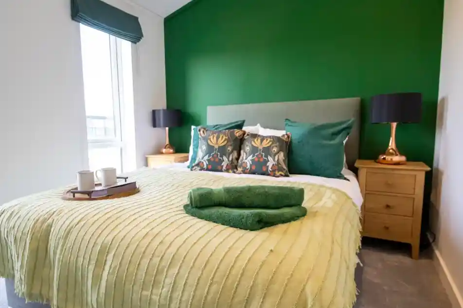 A brightly decorated bedroom with a large green accent wall behind a bed with green and patterned pillows, a light green blanket, and a tray with two mugs.