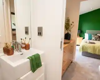 The image shows a modern, cozy en-suite bedroom and bathroom setup. In the foreground, there’s a compact white sink with a chrome faucet, a green towel draped neatly over the edge, and decorative amber-colored soap dispensers and a toothbrush holder with bamboo toothbrushes. A mirror above the sink reflects part of the bathroom.

Through the open door, a dresser with natural wood drawers and a decorative vase with greenery is visible. Beyond that, the bedroom features a large bed with a green accent wall, a grey upholstered headboard, and bedding in shades of green and white, along with decorative pillows. There are bedside tables on either side, one with a lamp. The overall look is fresh, stylish, and inviting, with green tones tying the two spaces together.