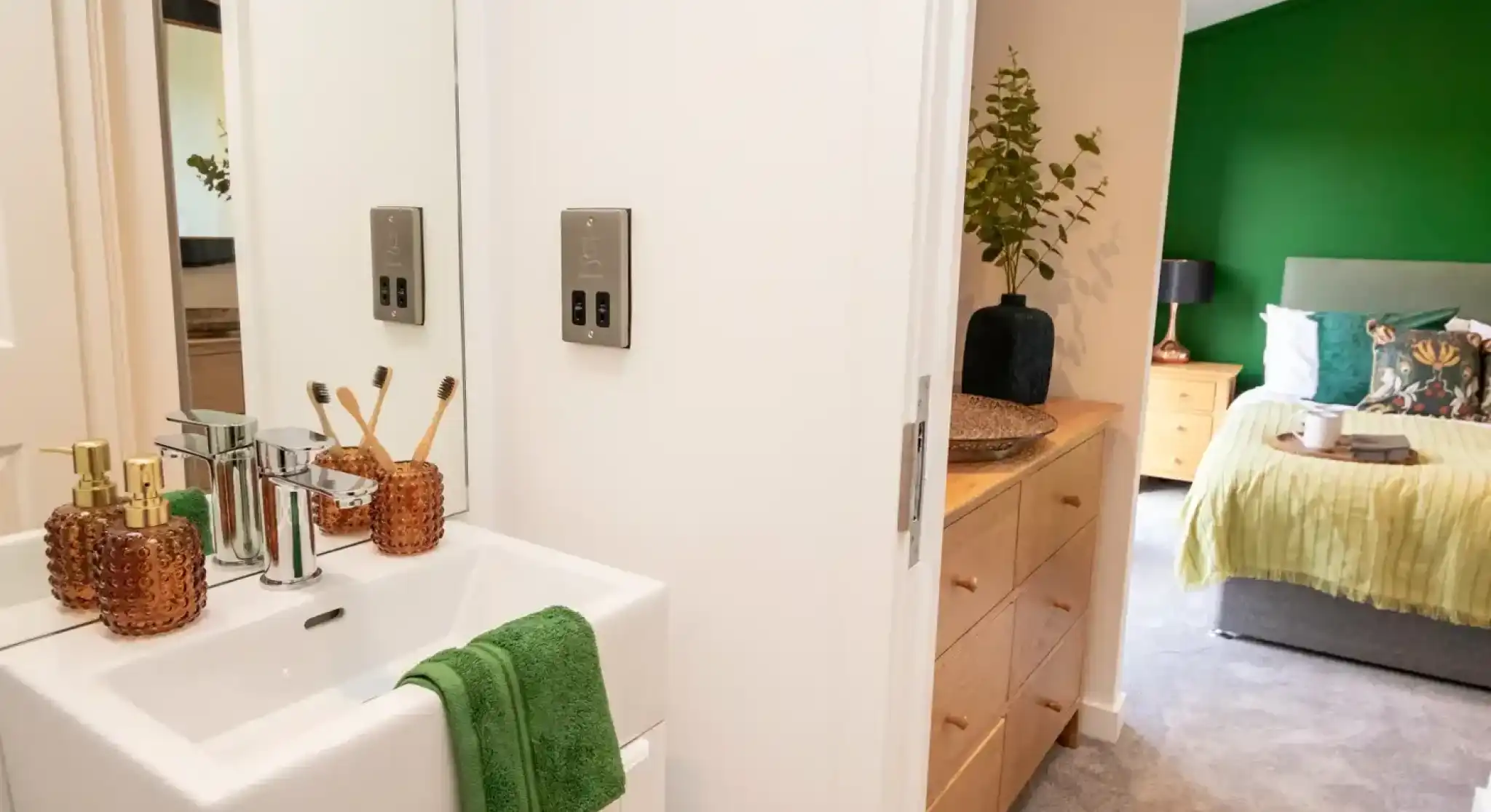 The image shows a modern, cozy en-suite bedroom and bathroom setup. In the foreground, there’s a compact white sink with a chrome faucet, a green towel draped neatly over the edge, and decorative amber-colored soap dispensers and a toothbrush holder with bamboo toothbrushes. A mirror above the sink reflects part of the bathroom.

Through the open door, a dresser with natural wood drawers and a decorative vase with greenery is visible. Beyond that, the bedroom features a large bed with a green accent wall, a grey upholstered headboard, and bedding in shades of green and white, along with decorative pillows. There are bedside tables on either side, one with a lamp. The overall look is fresh, stylish, and inviting, with green tones tying the two spaces together.