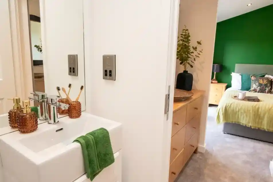 The image shows a modern, cozy en-suite bedroom and bathroom setup. In the foreground, there’s a compact white sink with a chrome faucet, a green towel draped neatly over the edge, and decorative amber-colored soap dispensers and a toothbrush holder with bamboo toothbrushes. A mirror above the sink reflects part of the bathroom.

Through the open door, a dresser with natural wood drawers and a decorative vase with greenery is visible. Beyond that, the bedroom features a large bed with a green accent wall, a grey upholstered headboard, and bedding in shades of green and white, along with decorative pillows. There are bedside tables on either side, one with a lamp. The overall look is fresh, stylish, and inviting, with green tones tying the two spaces together.