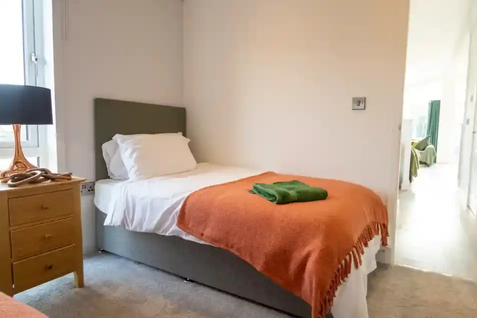 Twin bedroom with a single bed dressed in white bedding and an orange throw, a green towel folded on top, a wooden nightstand with a lamp by the window, and a view through the doorway into another room.