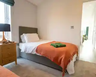 Twin bedroom with a single bed dressed in white bedding and an orange throw, a green towel folded on top, a wooden nightstand with a lamp by the window, and a view through the doorway into another room.
