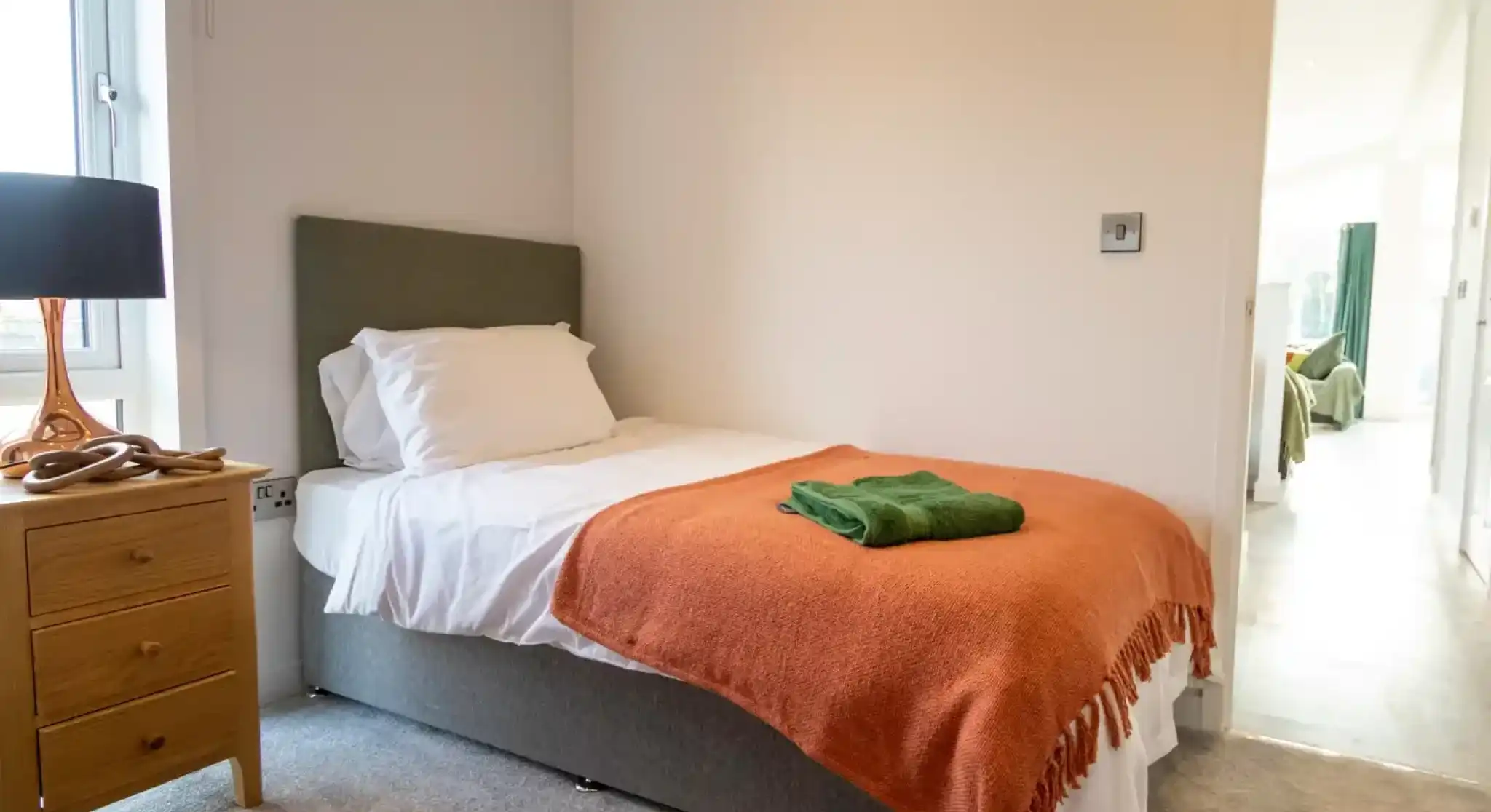 Twin bedroom with a single bed dressed in white bedding and an orange throw, a green towel folded on top, a wooden nightstand with a lamp by the window, and a view through the doorway into another room.