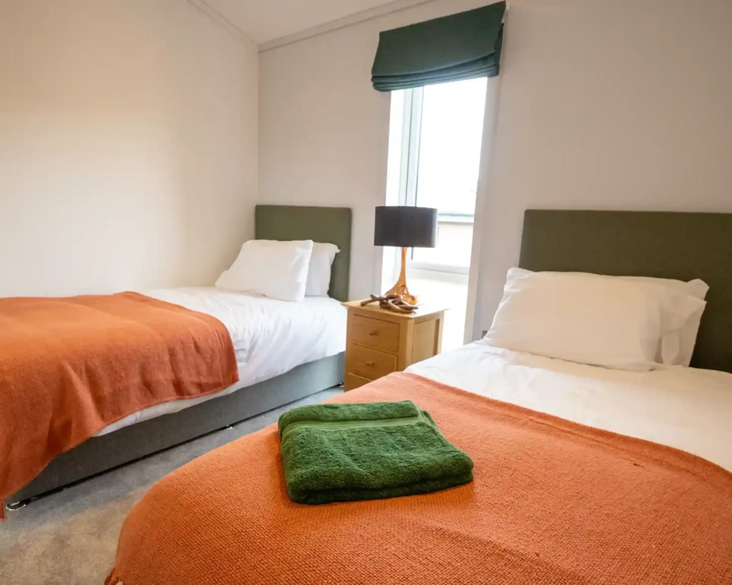 Twin bedroom with two single beds featuring white bedding and orange throws, a wooden nightstand with a lamp between them, and a window with a green roman shade.
