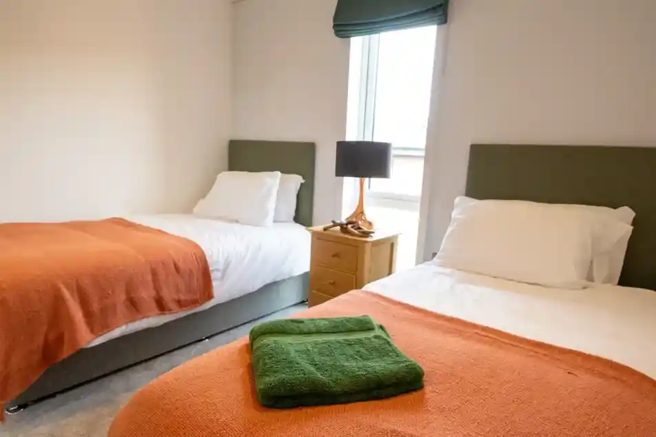 Twin bedroom with two single beds featuring white bedding and orange throws, a wooden nightstand with a lamp between them, and a window with a green roman shade.