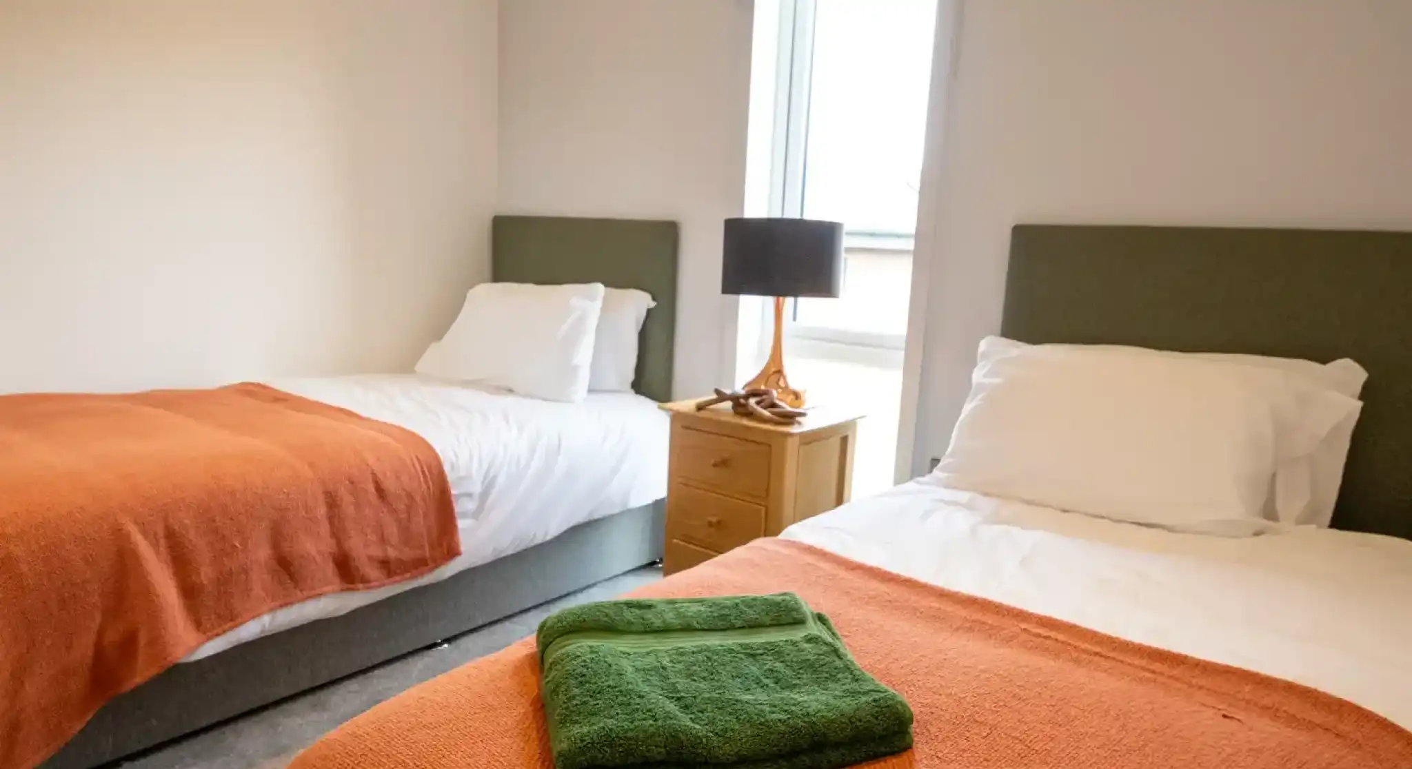 Twin bedroom with two single beds featuring white bedding and orange throws, a wooden nightstand with a lamp between them, and a window with a green roman shade.
