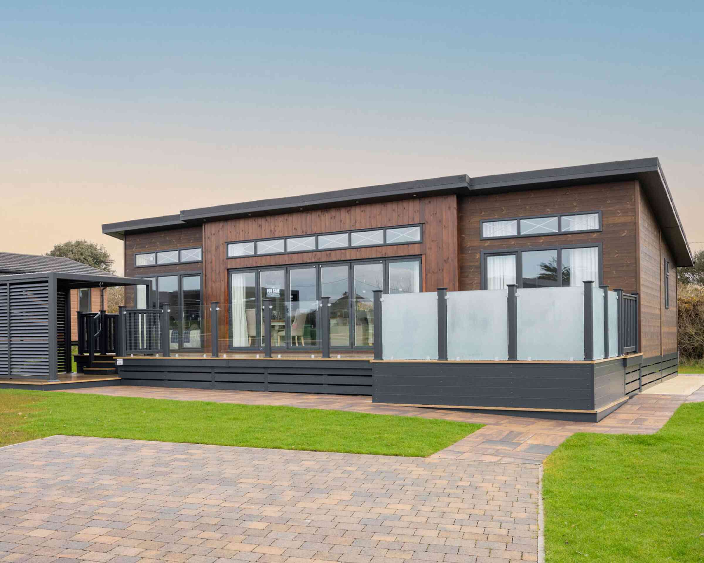 This image shows the exterior of a modern, single-story lodge-style home with a contemporary design. The structure features dark wood cladding with large floor-to-ceiling windows, allowing for ample natural light inside. The roof has a sleek, flat design with extended eaves, and narrow transom windows are positioned above the main windows for additional light and style. A spacious raised deck with frosted glass panel railings extends from the front, providing an outdoor living area. The surrounding landscape is well-maintained, with green grass, paved walkways, and a driveway made of interlocking bricks. The overall appearance is clean, stylish, and well-integrated with its natural surroundings.