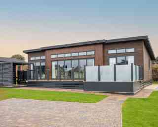 This image shows the exterior of a modern, single-story lodge-style home with a contemporary design. The structure features dark wood cladding with large floor-to-ceiling windows, allowing for ample natural light inside. The roof has a sleek, flat design with extended eaves, and narrow transom windows are positioned above the main windows for additional light and style. A spacious raised deck with frosted glass panel railings extends from the front, providing an outdoor living area. The surrounding landscape is well-maintained, with green grass, paved walkways, and a driveway made of interlocking bricks. The overall appearance is clean, stylish, and well-integrated with its natural surroundings.
