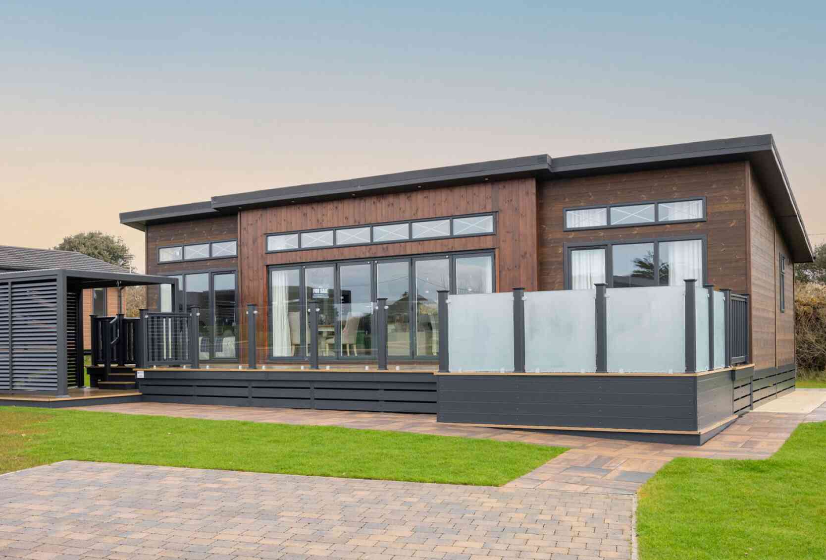 This image shows the exterior of a modern, single-story lodge-style home with a contemporary design. The structure features dark wood cladding with large floor-to-ceiling windows, allowing for ample natural light inside. The roof has a sleek, flat design with extended eaves, and narrow transom windows are positioned above the main windows for additional light and style. A spacious raised deck with frosted glass panel railings extends from the front, providing an outdoor living area. The surrounding landscape is well-maintained, with green grass, paved walkways, and a driveway made of interlocking bricks. The overall appearance is clean, stylish, and well-integrated with its natural surroundings.