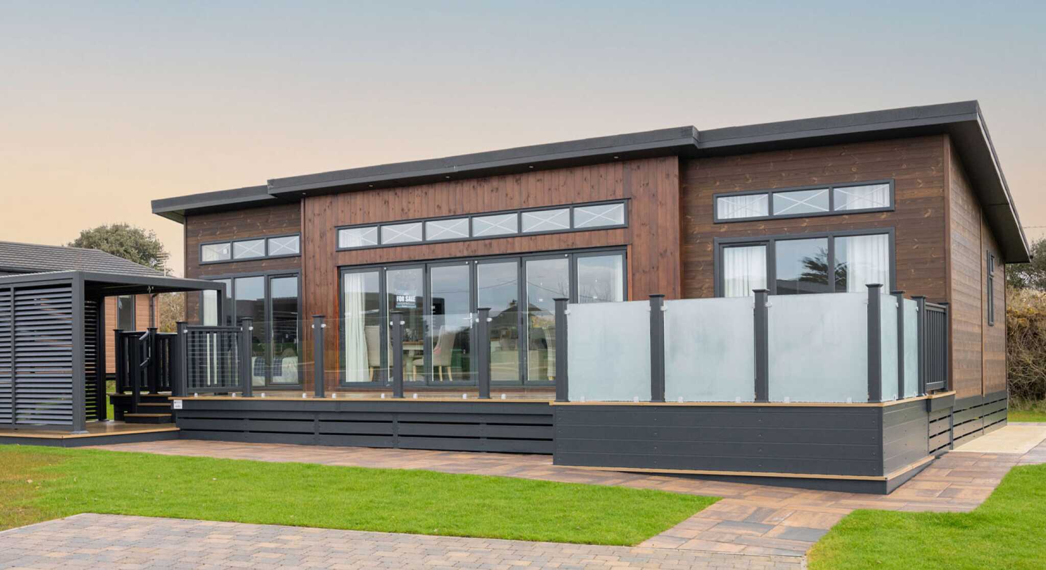 This image shows the exterior of a modern, single-story lodge-style home with a contemporary design. The structure features dark wood cladding with large floor-to-ceiling windows, allowing for ample natural light inside. The roof has a sleek, flat design with extended eaves, and narrow transom windows are positioned above the main windows for additional light and style. A spacious raised deck with frosted glass panel railings extends from the front, providing an outdoor living area. The surrounding landscape is well-maintained, with green grass, paved walkways, and a driveway made of interlocking bricks. The overall appearance is clean, stylish, and well-integrated with its natural surroundings.