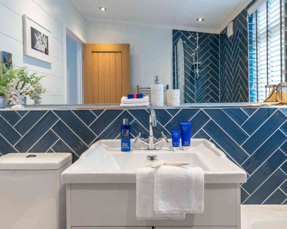 Modern bathroom with a herringbone-patterned blue tiled wall. A white sink with a sleek faucet is displayed, topped with a towel and blue skincare products. Natural light illuminates the space, highlighting the wooden door and decorative elements.