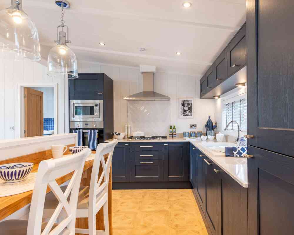 Modern kitchen featuring dark blue cabinetry, a stainless steel range hood, and a spacious countertop. A wooden dining table with white chairs is visible, and decorative items are neatly arranged on the counter. The space is well-lit with pendant lights above the table and windows allowing natural light.
