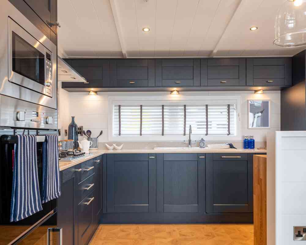 Modern kitchen featuring dark navy blue cabinetry, silver hardware, and a light countertop. Equipped with built-in appliances, including a microwave and oven. Natural light streams in through a window with white shutters, illuminating the space.
