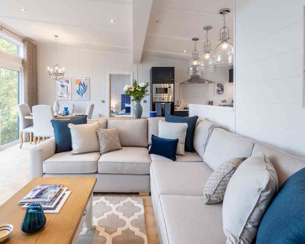 A modern living room with a large light-colored sectional sofa adorned with decorative pillows in various shades of blue and gray. A wooden coffee table sits in front, featuring magazines and a blue decorative item. The space includes stylish light fixtures, a dining area in the background, and large windows allowing natural light to fill the room.