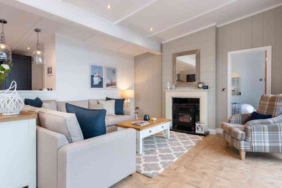 This image depicts a cosy and stylish living room with a soft, neutral colour palette accented by navy blue details. A large L-shaped beige sofa with a mix of textured and coloured cushions anchors the space, facing a white coffee table with a wooden top. To the right, a comfortable armchair with a plaid pattern adds a traditional touch. A modern electric fireplace is set into a light grey panelled wall, topped with a wide wooden-framed mirror. Above the sofa, minimalist art prints enhance the contemporary feel, while pendant lights from the adjoining kitchen add ambient light. The room features light wood flooring with a patterned area rug, creating a warm and welcoming atmosphere. A glimpse into a neatly made bedroom is visible through a door to the right, adding to the open, homely feel of the space.