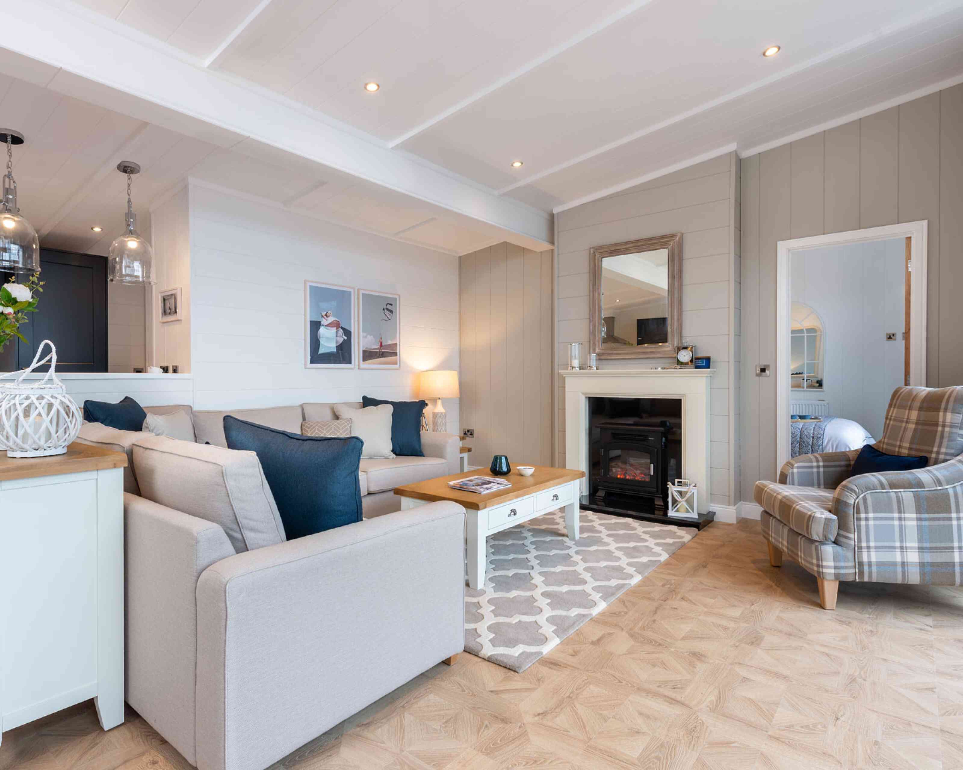 This image depicts a cosy and stylish living room with a soft, neutral colour palette accented by navy blue details. A large L-shaped beige sofa with a mix of textured and coloured cushions anchors the space, facing a white coffee table with a wooden top. To the right, a comfortable armchair with a plaid pattern adds a traditional touch. A modern electric fireplace is set into a light grey panelled wall, topped with a wide wooden-framed mirror. Above the sofa, minimalist art prints enhance the contemporary feel, while pendant lights from the adjoining kitchen add ambient light. The room features light wood flooring with a patterned area rug, creating a warm and welcoming atmosphere. A glimpse into a neatly made bedroom is visible through a door to the right, adding to the open, homely feel of the space.