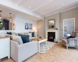 This image depicts a cosy and stylish living room with a soft, neutral colour palette accented by navy blue details. A large L-shaped beige sofa with a mix of textured and coloured cushions anchors the space, facing a white coffee table with a wooden top. To the right, a comfortable armchair with a plaid pattern adds a traditional touch. A modern electric fireplace is set into a light grey panelled wall, topped with a wide wooden-framed mirror. Above the sofa, minimalist art prints enhance the contemporary feel, while pendant lights from the adjoining kitchen add ambient light. The room features light wood flooring with a patterned area rug, creating a warm and welcoming atmosphere. A glimpse into a neatly made bedroom is visible through a door to the right, adding to the open, homely feel of the space.