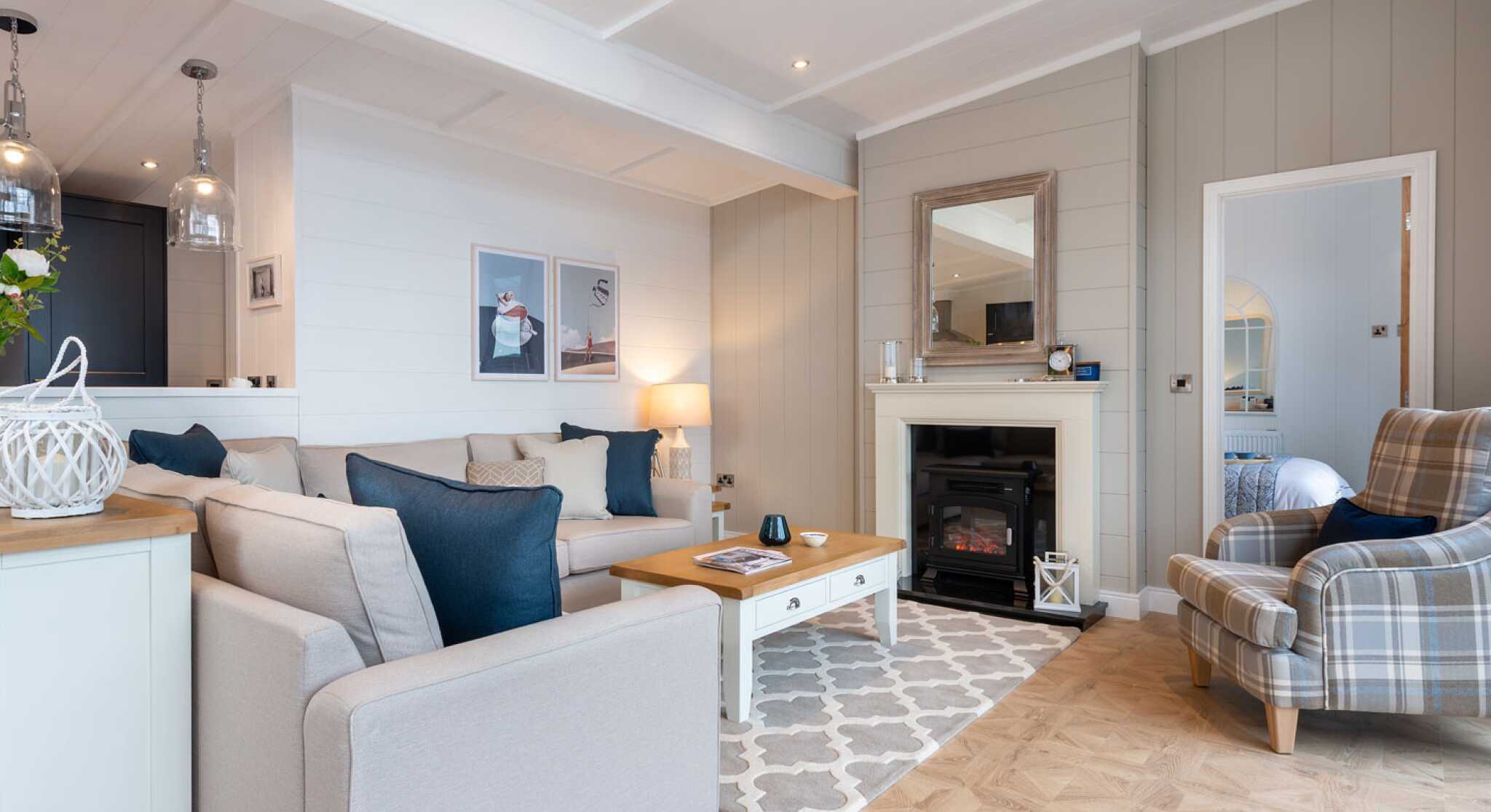 This image depicts a cosy and stylish living room with a soft, neutral colour palette accented by navy blue details. A large L-shaped beige sofa with a mix of textured and coloured cushions anchors the space, facing a white coffee table with a wooden top. To the right, a comfortable armchair with a plaid pattern adds a traditional touch. A modern electric fireplace is set into a light grey panelled wall, topped with a wide wooden-framed mirror. Above the sofa, minimalist art prints enhance the contemporary feel, while pendant lights from the adjoining kitchen add ambient light. The room features light wood flooring with a patterned area rug, creating a warm and welcoming atmosphere. A glimpse into a neatly made bedroom is visible through a door to the right, adding to the open, homely feel of the space.