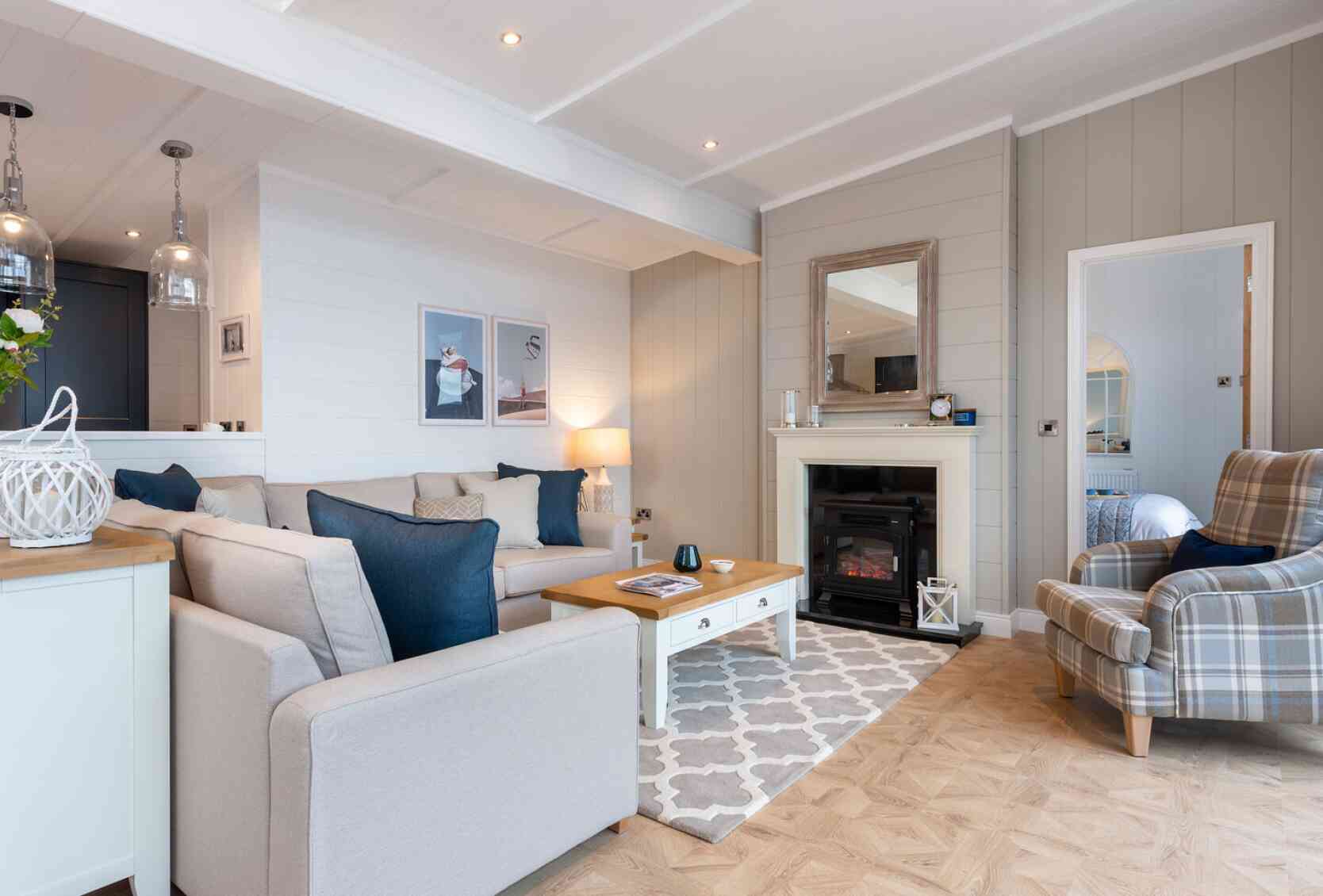 This image depicts a cosy and stylish living room with a soft, neutral colour palette accented by navy blue details. A large L-shaped beige sofa with a mix of textured and coloured cushions anchors the space, facing a white coffee table with a wooden top. To the right, a comfortable armchair with a plaid pattern adds a traditional touch. A modern electric fireplace is set into a light grey panelled wall, topped with a wide wooden-framed mirror. Above the sofa, minimalist art prints enhance the contemporary feel, while pendant lights from the adjoining kitchen add ambient light. The room features light wood flooring with a patterned area rug, creating a warm and welcoming atmosphere. A glimpse into a neatly made bedroom is visible through a door to the right, adding to the open, homely feel of the space.