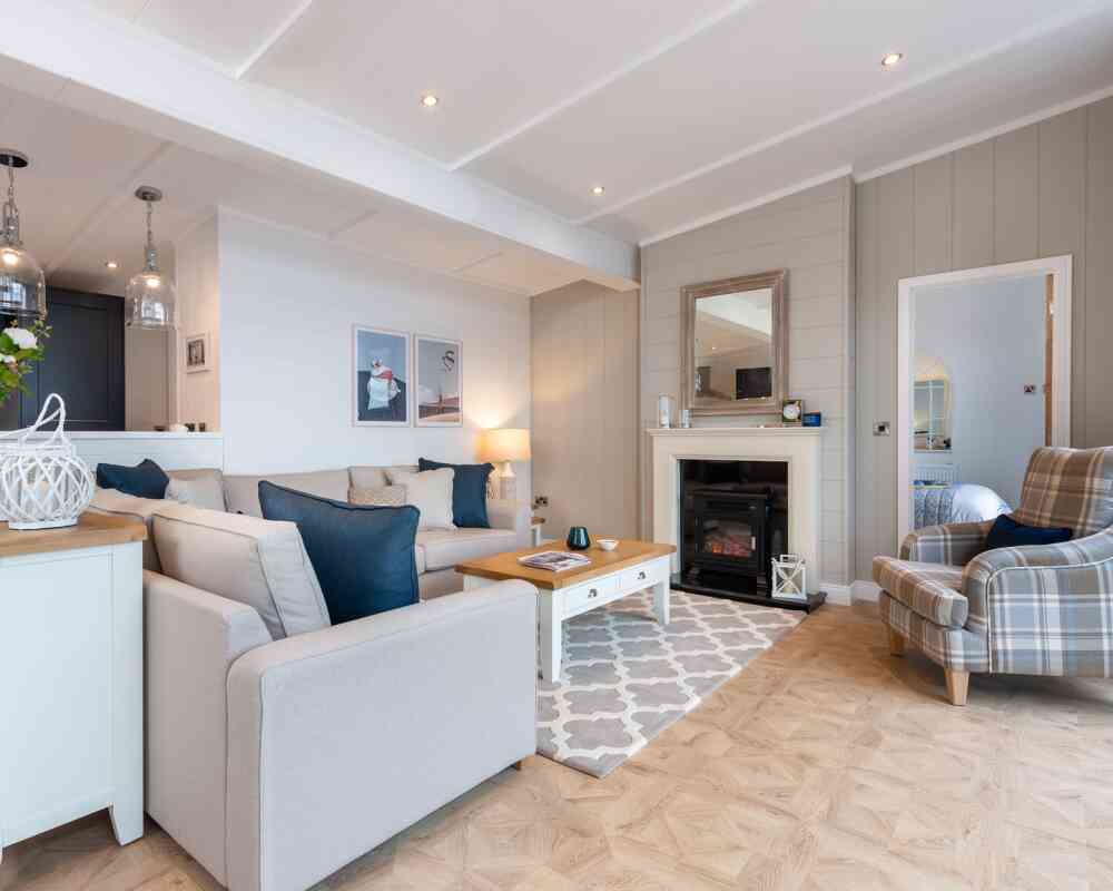 This image depicts a cosy and stylish living room with a soft, neutral colour palette accented by navy blue details. A large L-shaped beige sofa with a mix of textured and coloured cushions anchors the space, facing a white coffee table with a wooden top. To the right, a comfortable armchair with a plaid pattern adds a traditional touch. A modern electric fireplace is set into a light grey panelled wall, topped with a wide wooden-framed mirror. Above the sofa, minimalist art prints enhance the contemporary feel, while pendant lights from the adjoining kitchen add ambient light. The room features light wood flooring with a patterned area rug, creating a warm and welcoming atmosphere. A glimpse into a neatly made bedroom is visible through a door to the right, adding to the open, homely feel of the space.