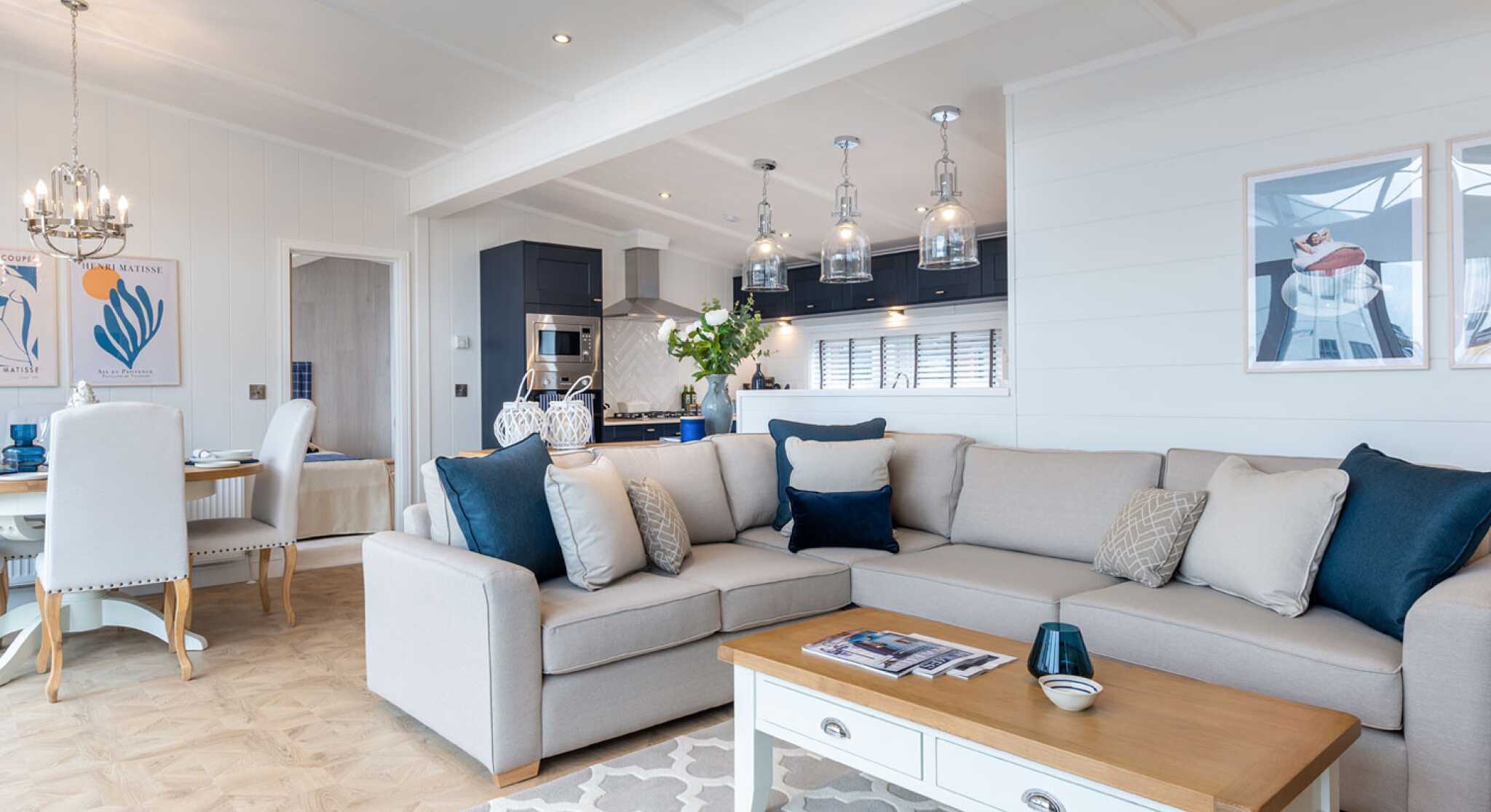 This image shows a bright and modern open-plan living space that combines a lounge, dining area, and kitchen. The interior features a soft colour palette with whites, greys, and navy blue accents. A large beige sectional sofa with various throw pillows sits on a patterned rug, facing a white coffee table with a wooden top. Above the kitchen island hang four glass pendant lights, and the kitchen itself has dark cabinetry with stainless steel appliances. The dining area has a round wooden table surrounded by white upholstered chairs. The walls are adorned with contemporary artwork, and the space is well-lit with recessed ceiling lights and natural light coming through large windows. The overall décor is coastal and elegant, giving a relaxed yet upscale ambiance.