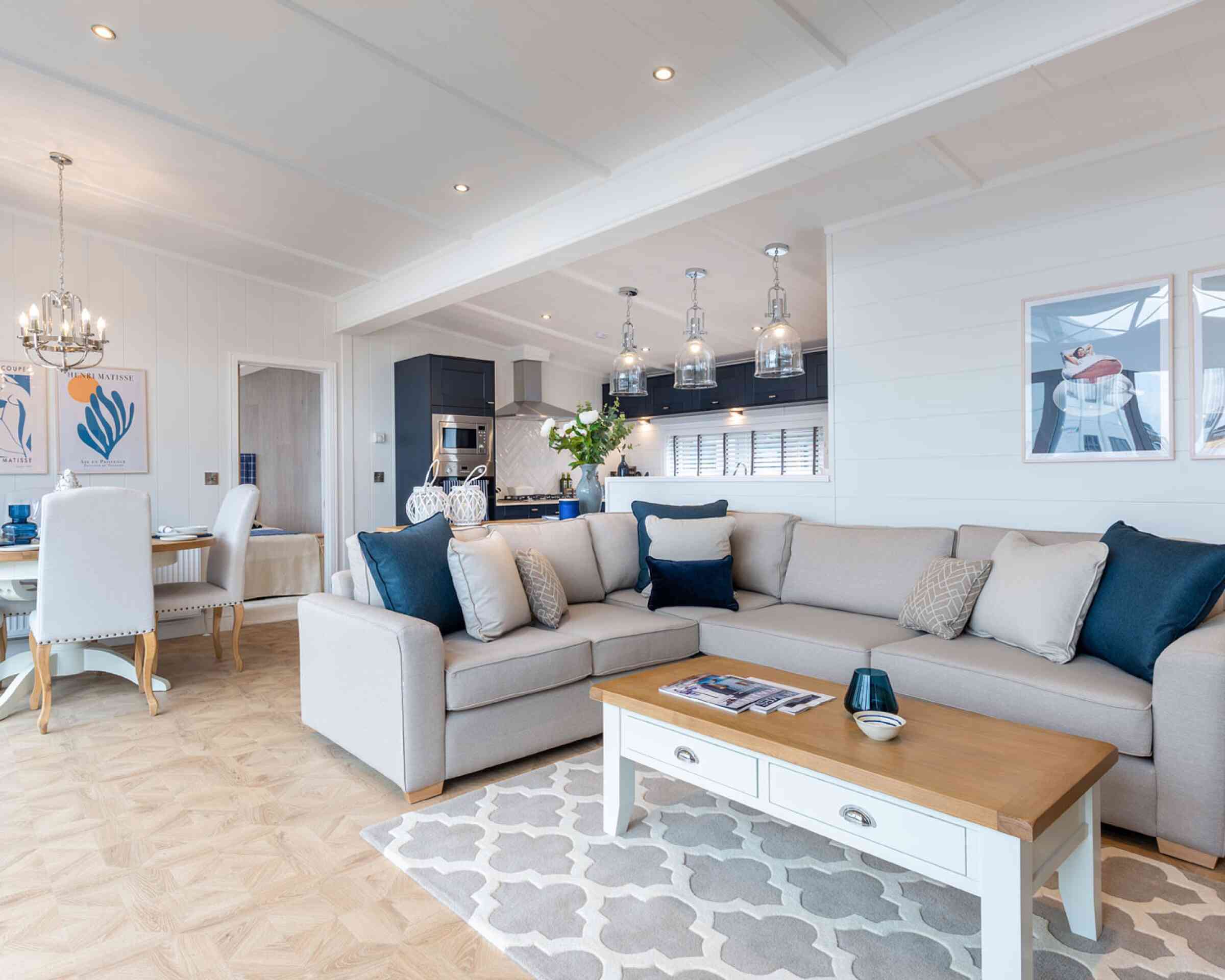 This image shows a bright and modern open-plan living space that combines a lounge, dining area, and kitchen. The interior features a soft colour palette with whites, greys, and navy blue accents. A large beige sectional sofa with various throw pillows sits on a patterned rug, facing a white coffee table with a wooden top. Above the kitchen island hang four glass pendant lights, and the kitchen itself has dark cabinetry with stainless steel appliances. The dining area has a round wooden table surrounded by white upholstered chairs. The walls are adorned with contemporary artwork, and the space is well-lit with recessed ceiling lights and natural light coming through large windows. The overall décor is coastal and elegant, giving a relaxed yet upscale ambiance.