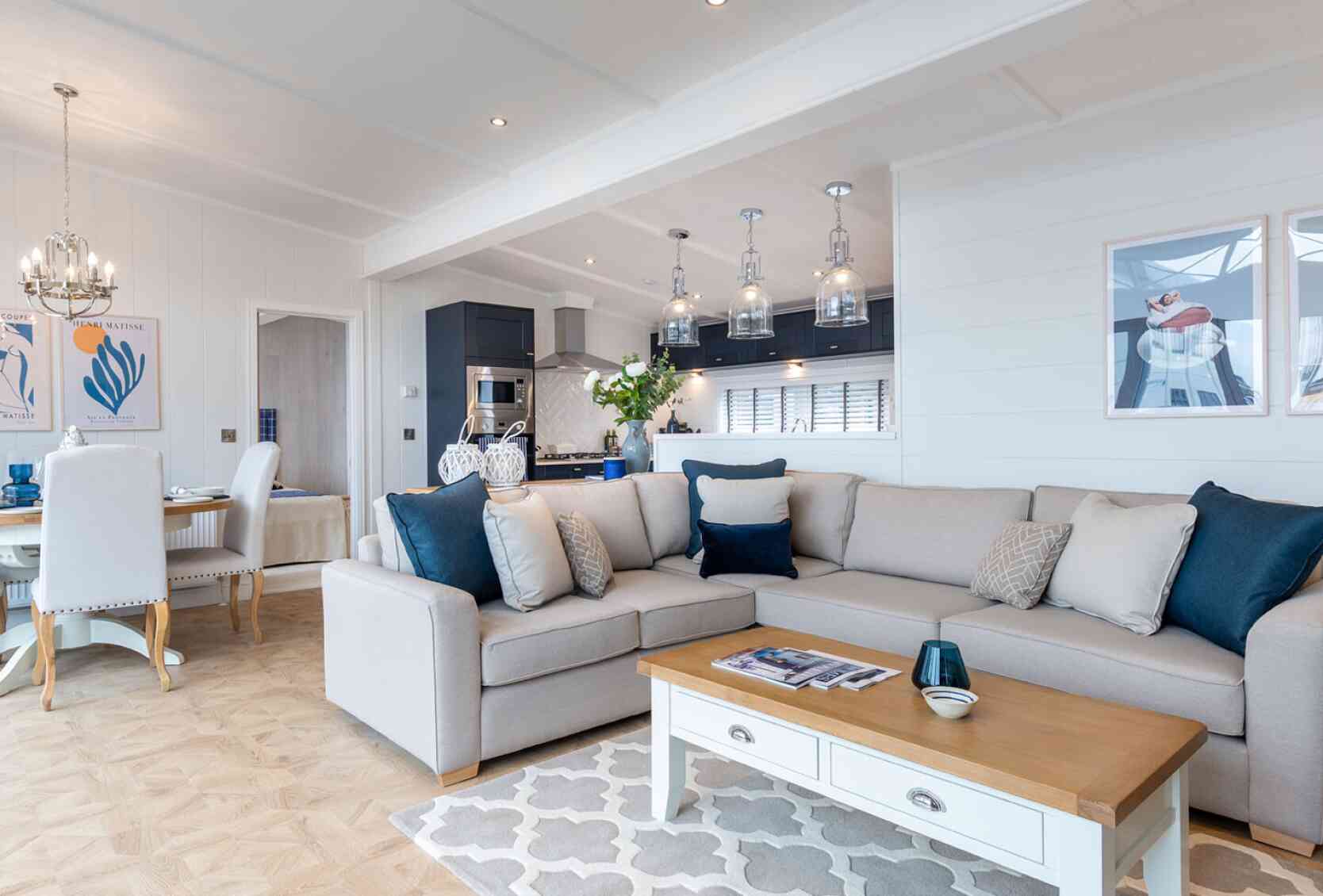 This image shows a bright and modern open-plan living space that combines a lounge, dining area, and kitchen. The interior features a soft colour palette with whites, greys, and navy blue accents. A large beige sectional sofa with various throw pillows sits on a patterned rug, facing a white coffee table with a wooden top. Above the kitchen island hang four glass pendant lights, and the kitchen itself has dark cabinetry with stainless steel appliances. The dining area has a round wooden table surrounded by white upholstered chairs. The walls are adorned with contemporary artwork, and the space is well-lit with recessed ceiling lights and natural light coming through large windows. The overall décor is coastal and elegant, giving a relaxed yet upscale ambiance.