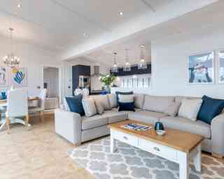This image shows a bright and modern open-plan living space that combines a lounge, dining area, and kitchen. The interior features a soft colour palette with whites, greys, and navy blue accents. A large beige sectional sofa with various throw pillows sits on a patterned rug, facing a white coffee table with a wooden top. Above the kitchen island hang four glass pendant lights, and the kitchen itself has dark cabinetry with stainless steel appliances. The dining area has a round wooden table surrounded by white upholstered chairs. The walls are adorned with contemporary artwork, and the space is well-lit with recessed ceiling lights and natural light coming through large windows. The overall décor is coastal and elegant, giving a relaxed yet upscale ambiance.