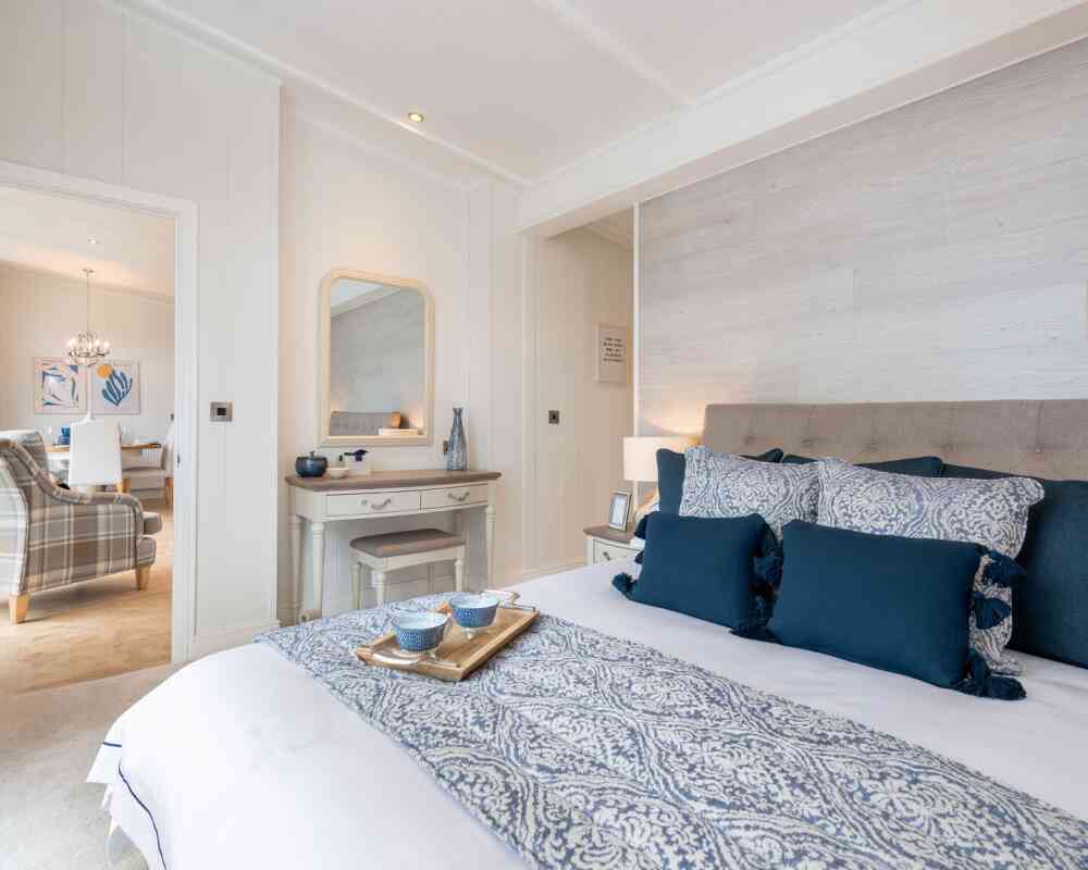 A cozy bedroom featuring a neatly made bed with blue and white patterned bedding and decorative pillows. A wooden dresser with a mirror is nearby, and a living area can be seen in the background with a chandelier and plaid sofa. Soft lighting enhances the inviting atmosphere.